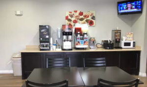 Breakfast display counter with tea, coffee, juice dispensers, microwave and waffle machine, wall mounted art, tables, chairs, wall mounted TV