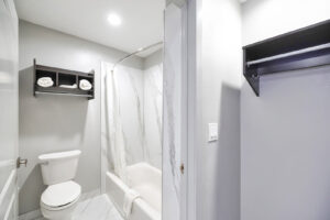 Shower tub with shower curtain and bathmat, toilet, unit with towels, tiled flooring, shelf with hanging rail