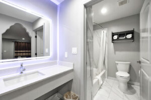 Shower tub with shower curtain and bathmat, toilet, unit with towels, vanity unit, back lit mirror, tiled flooring