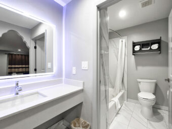 Shower tub with shower curtain and bathmat, toilet, unit with towels, vanity unit, back lit mirror, tiled flooring