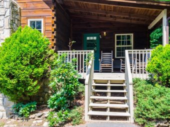 Cabin exterior showing wooden steps with small bushes leading to entrance door and porch area with rocking shairs