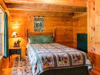 Wood panelled bedroom with double bed, night stand with decorative lamp, wooden drawers unit, wooden floor with large rug and wall mounted tapestry