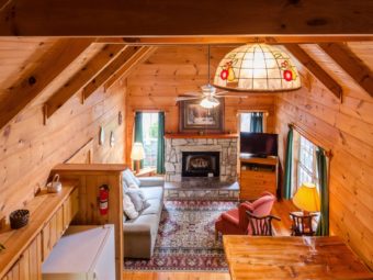 Cabin interior viewed from loft bedroom area with decorative ceiling lampshade and fire extinguisher looking down on sitting area with sofa, easy chair, side table with lap, fireplace, corner wooden storage unit with flat screen TV, wooden flooring with large rug