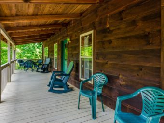 Tree shaded wooden porch with patio dining set, rocking chairs and extra chairs, cabin door and exterior light.