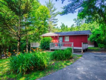 Red painted cottage exterior showing wooden deck with wooden railings, trees, landscaping and parking area