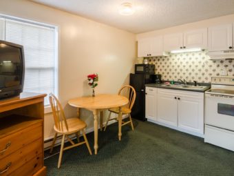 Wooden storage unit with TV, wooden table and two chairs, fridge, microwave, kitchen wall cabinets, double sink, stove and carpet flooring