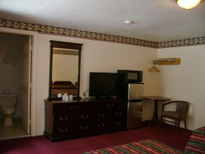 toilet in separate room, large wall mounted mirror, wooden units with drawers, flat screen tv, fridge, microwave, small table, upholstered chair, hanging rail with hangers and overhead shelf, edge of two queen beds and carpet flooring