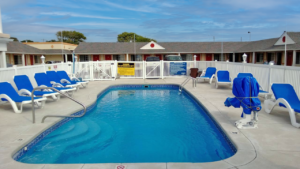 Outdoor swimming pool with ADA approved pool lift. Pool is enclosed by a safety fencing and entrance gate, concrete pool surround with patio chairs