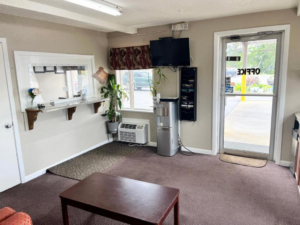 Guest check-in desk, plants, water dispenser, occasional table, wall mounted TV, door to exterior parking, carpet flooring