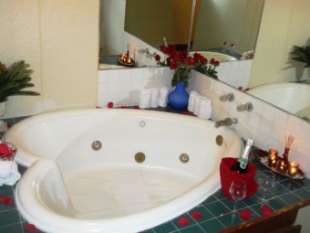 spa tub, mirrored walls, tiled surround, bottle of champagne, wine glasses, vase with roses, candles and incense sticks on a tray, small green plant in vase