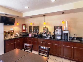 Breakfast counter display with water dispenser, coffee machines, waffle iron, toaster, disposable plates and silver ware, cereal dispensers, fridge, tables and chairs and tiled flooring