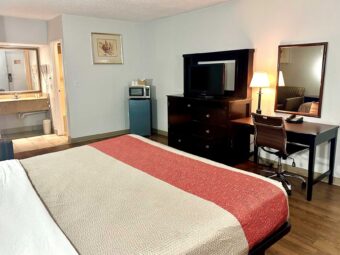 King bed, sofa, fridge with microwave, wooden drawer unit with TV, desk with lamp, chair and telephone, mirror, art image, laminate flooring, alcove with vanity unit, mirror and hair dryer, doorway to bathroom, tiled flooring