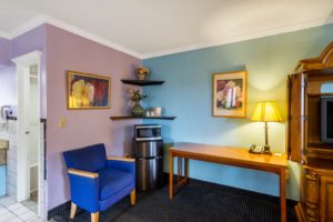 Wooden console with tv, desk with lamp and telephone, wall mounted art, fridge, microwave, corner shelves above with ice bucket and vases containing flowers, easy chair, doorway to bathroom and carpet flooring