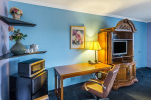 Corner shelves with vases containing flowers, ice bucket, fridge, microwavem desk with office chair, lamp and telephone, wall mounted art, woodem console with tv and carpet flooring