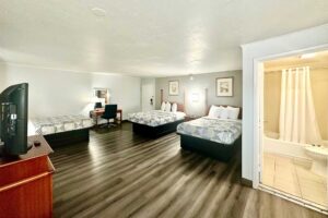 Three double beds, art images, desk with chair, mirror, unit with TV, laminate flooring, doorway to bathroom with shower tub, toilet, tiled flooring