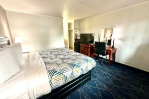 King bed, night stand with bedside lamp. desk with chair and lamp, mirror, drawer unit with TV, fridge with microwave, ironing board and iron, alcove with hanging rail, carpet flooring