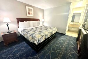King bed, night stands with bedside lamps, art image, fridge with microwave, drawer unit with TV, carpet flooring, alcove with vanity unit and mirror, tiled flooring