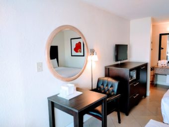 Wall mounted mirror, small table with ice bucket and upholstered chair, wall mounted light, wooden unit with drawers and microwave, wall mounted flat screen tv, alcove with vanity unit, towels, bathroom amenitied, large wall mounted mirror and portable folding luggage rack, edge of two queen beds and tiled flooring