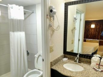 Large wall mounted mirror, wall mounted hair dryer, towels, bathroom amenities on vanity unit with sink, doorway to shower tub wuth shower curtain and toilet