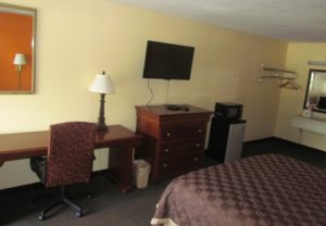 King bed, vanity unit, large wall mounted mirror with overhead strip light, wall mounted metal hanging rail with overhead shelf and hangers, microwave, fridge, wooden drawwer unit, wall mounted flat screen tv, desk with lamp and upholstered chair, wall mounted mirror and carpet flooring