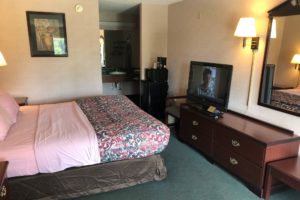 King bed. night stands, art image, fridge with microwave, low level wooden drawers with flatscreen TV, wall light, mirror, alcove with vanity unit, hanging rail with hangers, carpet flooring