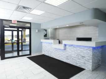 Guest check-in desk, large floor mat, two sets of double doors leading to hotel exterior, tiled flooring