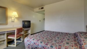 Queen bed, desk with chair and tv, large wall mounted mirror, wall mounted light and caret flooring