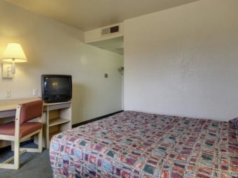 Queen bed, desk with chair and tv, large wall mounted mirror, wall mounted light and caret flooring