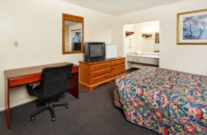 Queen bed, wall mounted art, wooden drawer unit with small fridge and tv, desk with chair, large wall mounted mirror, carpet flooring and alcove with vanity unit, large wall mounted mirror with overhead strip light and hanging rail with overhead shelf and wall mounted towel rack with towels