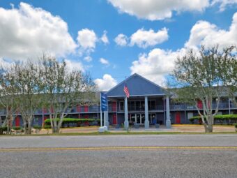 Two story building with canopied drive through entrance, exterior guest room entrances with covered walkway, grassy area with trees, American flag pole