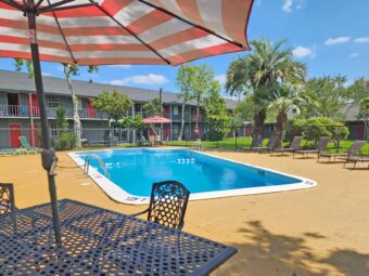 outdoor pool with fencing, patio tables with umbrella and chairs, sun loungers, two story hotel building overlooking pool with exterior guest room entrances