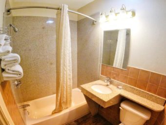 Shower tub with shower curtain, sink, wall mounted mirror with overhead lights, toilet, wall mounted towel rack with towels