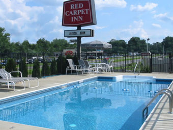 Outdoor pool, pool loungers and patio tables and chirs with parasol on concrete poolside, fencing and small trees