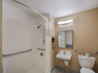 Shower Tub with grab handles and wall mounted towel rail with towel, sink, wall mounted mirroe with overhead strip light, wall mounted hair dryer, grab handles by toilet, bathroom amenities and tiled floor