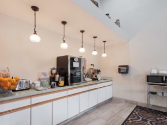 Breakfast counter with coffee machine, cereal dispenser, pitchers of juice, toaster, assorted breakfast pastries, fresh fruit , microwave, wall mounted paper towel dispenser, suspended lighting, disposable plates and silver ware, caarpet and tile flooring