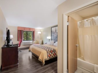 Shower tub with shower curtain in separate room, queen bed, large wall mounted art above bed, wall mounted bedside light, night stand with telephone, corner suspended light over easy chair, desk with chair on wheels, table lamp, microwave, wooden storage unit with drawers, flat screen TV and laminate flooring