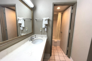 Vanity unit, mirror with overhead light, towl rail with towels, hairdryer, doorway to shower tub with shower curtain and bath mat, tiled flooring