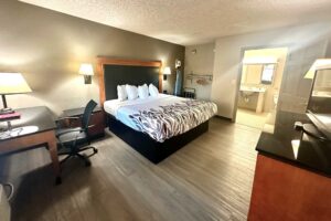 King bed, wall bedside lights, night stand, desk with lamp and chair, ironing board with iron, hanging rail with hangers, portable folding luggage rack, unit with TV, laminate flooring, doorway leading to the bathroom, vanity unit, mirror, toilet, tiled flooring