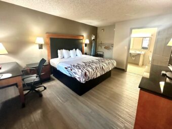 King bed, wall bedside lights, night stand, desk with lamp and chair, ironing board with iron, hanging rail with hangers, portable folding luggage rack, unit with TV, laminate flooring, doorway leading to the bathroom, vanity unit, mirror, toilet, tiled flooring