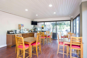 Breakfast counter with toaster, microwave, cereal dispenser, paper plates, bowls, plastic dinnerware, pastries display, self serve coffee pots, wall mounted TV, juice dispenser machine, table and chairs, laminate flooring