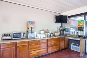 Breakfast counter with toaster, microwave, cereal dispenser, paper plates, bowls, plastic dinnerware, pastries display, self serve coffee pots, wall mounted TV, juice dispenser machine