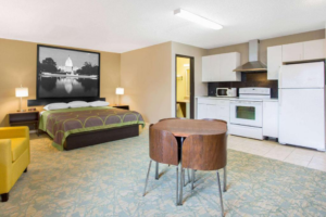 King bed, night stands with bedside lamps and clock, easy chair, table and chairs, kitchenette area with oven, microwave, fridge freezer and cabinets, doorway to bathroom, carpet and tiled flooring