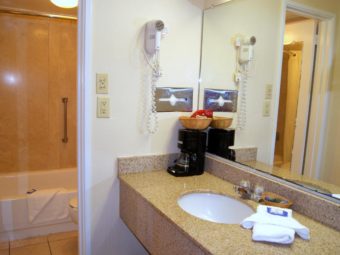 vanity unit with sink, coffee maker, towels, bathroom amenities, wall mounted tissue box, wall mounted hair dryer, large wall mounted mirror, doorway to bathroom, shower tub with bath mat and tiled flooring