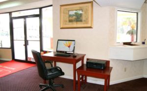 Desk with computer screen and keyboard and office chair, samll table with printer, wall mounted art, welcome mat and carpet flooring