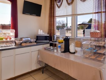 Breakfast counter display with fresh fruit, assorted breakfast pasties, toaster, juice pitcher, coffee machine, cereal dispenser, assorted snack bars, disposable plates and silverware, wall mounted flat screen tv and tiled flooring