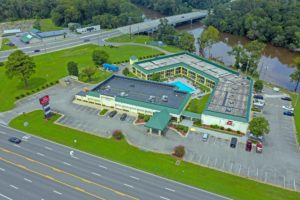 Drone view of hotel exterior, canopy hotel entrance,brand signage, parking spaces, two story building, outdoor pool, grassy areas, trees and small bushs, river with fishing desk