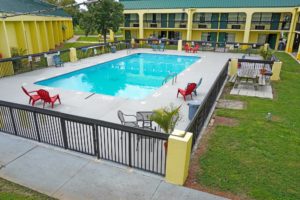 pool with concrete surround, pool chairs, table and chairs, safety fence, two story building with exterior room entrances, grassy areas