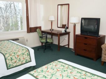 Two queen beds, desk with chair and lamp, wall mounted mirror, wooden drawer unit with tv, easy chair, carpet flooring
