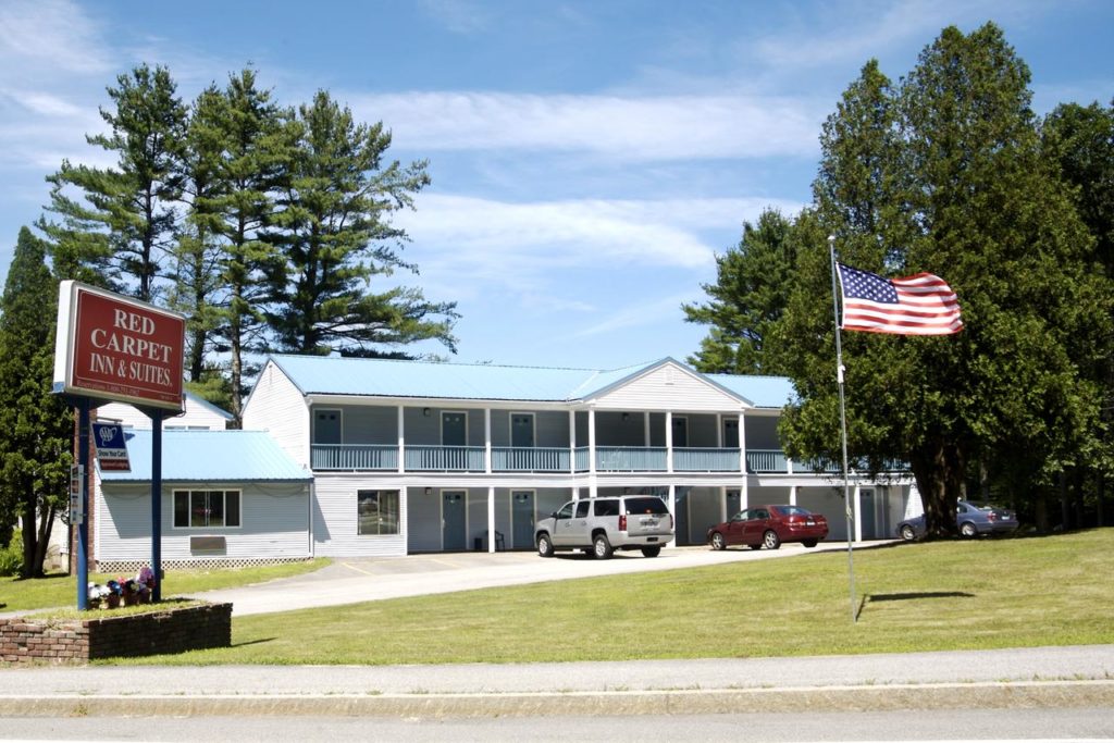 Hotel entrance, two storey building, parking spaces, grassy areas, american flag on pole, overlooked by tall trees