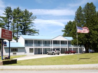 Hotel entrance, two storey building, parking spaces, grassy areas, american flag on pole, overlooked by tall trees